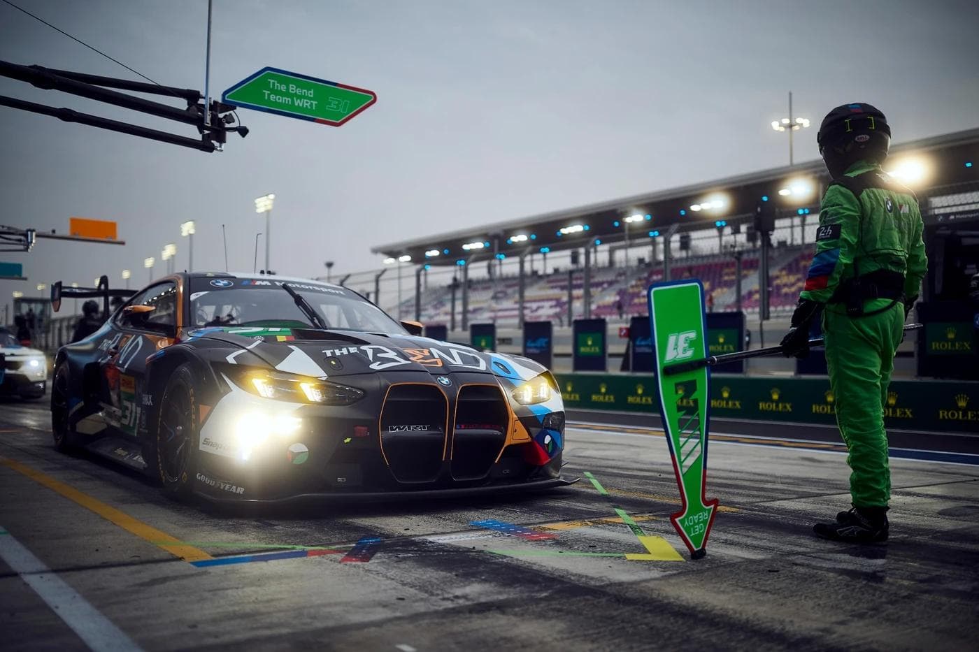 BMW M4 GT3 Evo number 31 entered by The Bend Team WRT in the pit lane at Lusail International Circuit during the 2025 FIA World Endurance Championship Qatar round, lollipop man in green race suit holding a get ready board at dusk