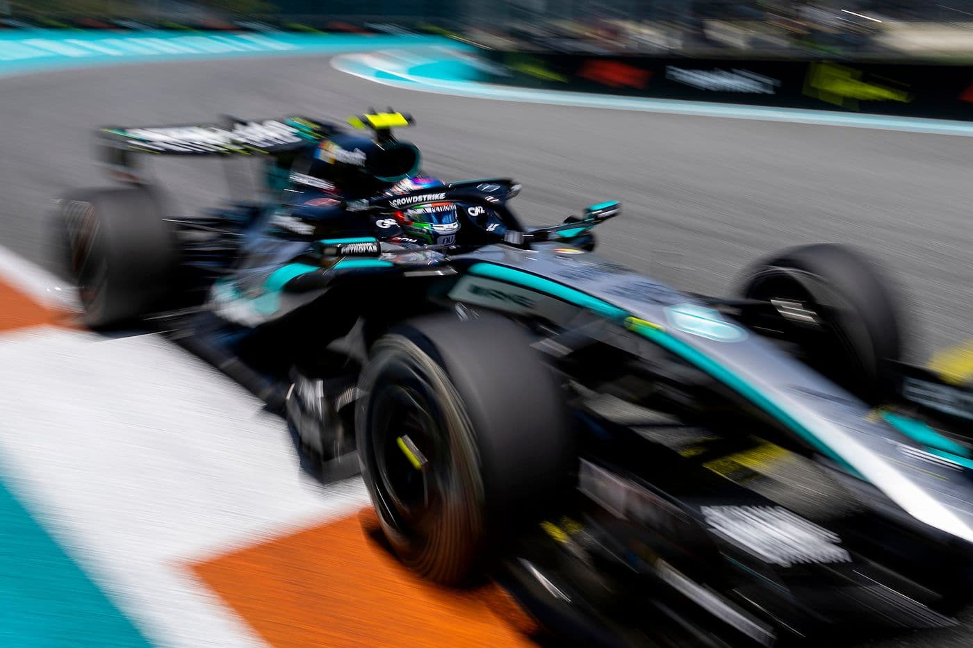 A Mercedes-AMG PETRONAS Formula 1 car speeds through a corner on a racetrack, captured with heavy motion blur that emphasises its rapid movement. The black and turquoise livery is visible, along with the blurred tyres and track markings in orange and white. The driver’s helmet and cockpit can be seen, though details are indistinct due to the high-speed motion.