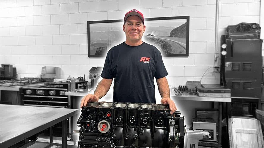 Jay Meagher, founder of RS Garage, standing proudly with the 2JZ RSG 1600 short block engine inside the workshop, with the engine and Jay in colour against a black-and-white background.