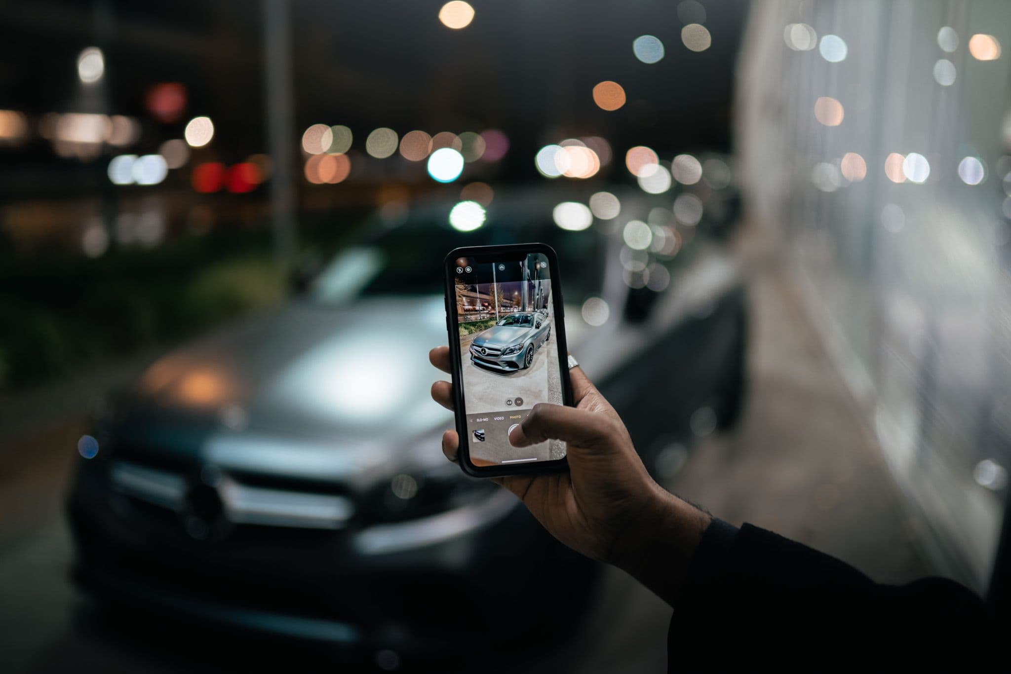 Hand holding a phone capturing a photo of a Mercedes-Benz parked outside at night.