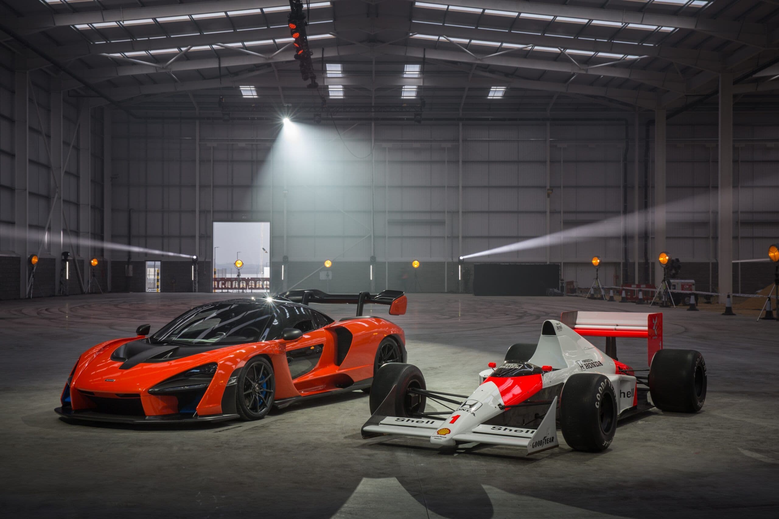 A McLaren Senna hypercar in vibrant orange sits next to a classic McLaren MP4/4 Formula 1 car in a dimly lit industrial warehouse, with dramatic overhead lighting casting reflections on the polished concrete floor.