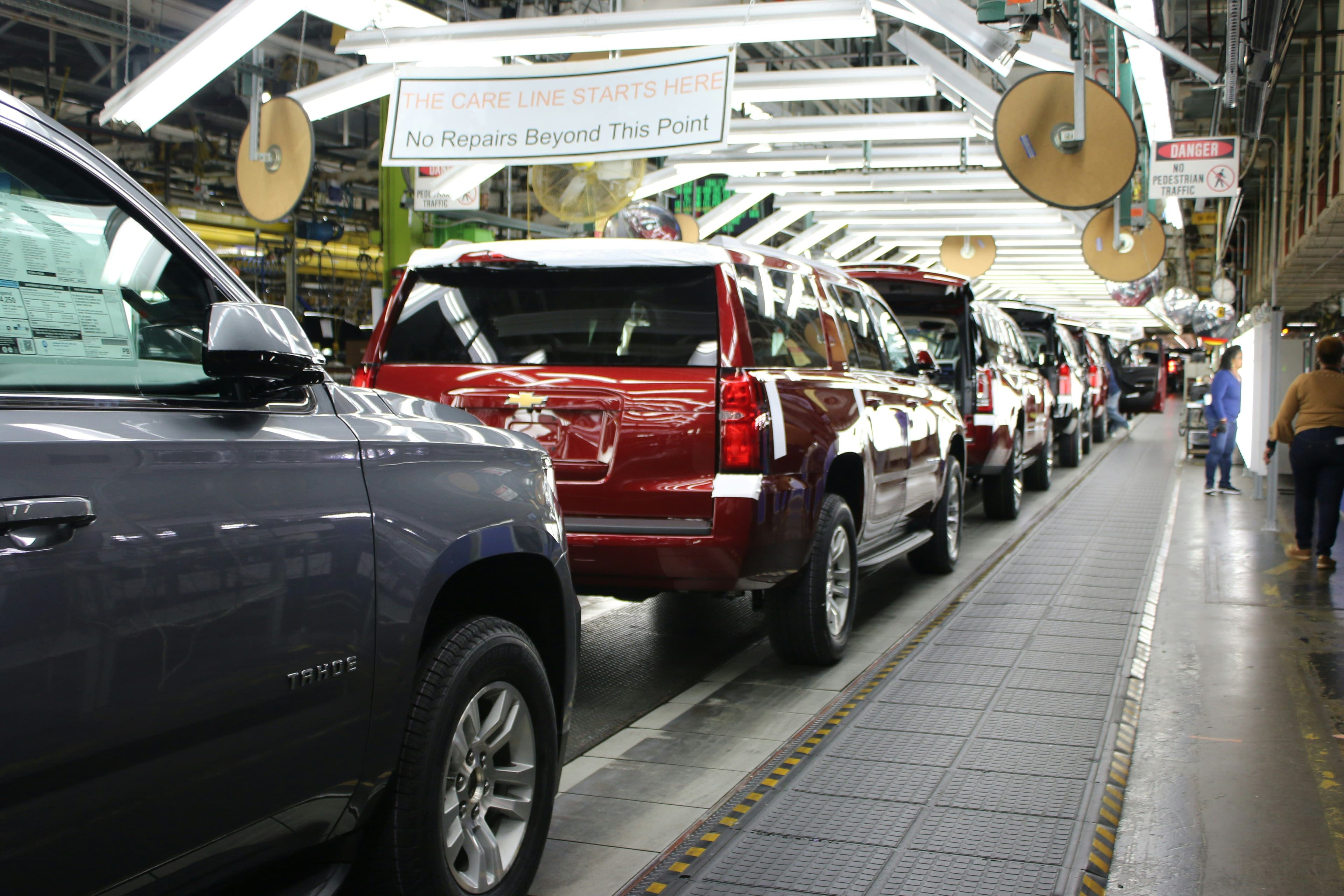 A Chevrolet SUV production line inside an automotive assembly plant. Several newly manufactured vehicles, including a red Chevrolet Suburban and a grey Chevrolet Tahoe, are lined up under bright overhead lights. Workers in the background inspect vehicles as they move along the final stages of assembly. A sign overhead reads, “The care line starts here – No repairs beyond this point.”