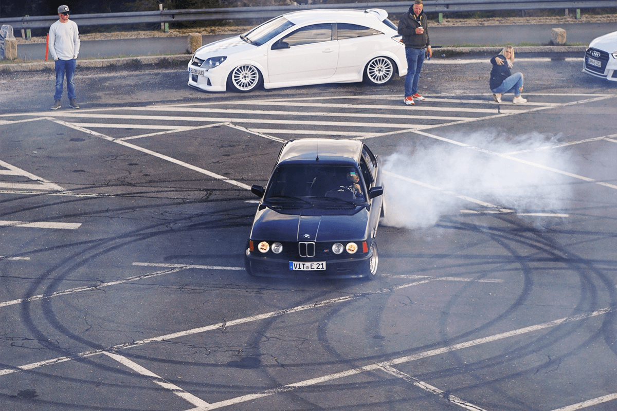 BMW E21 performing a burnout at Worthersee with smoke coming from the rear tyres on marked asphalt.