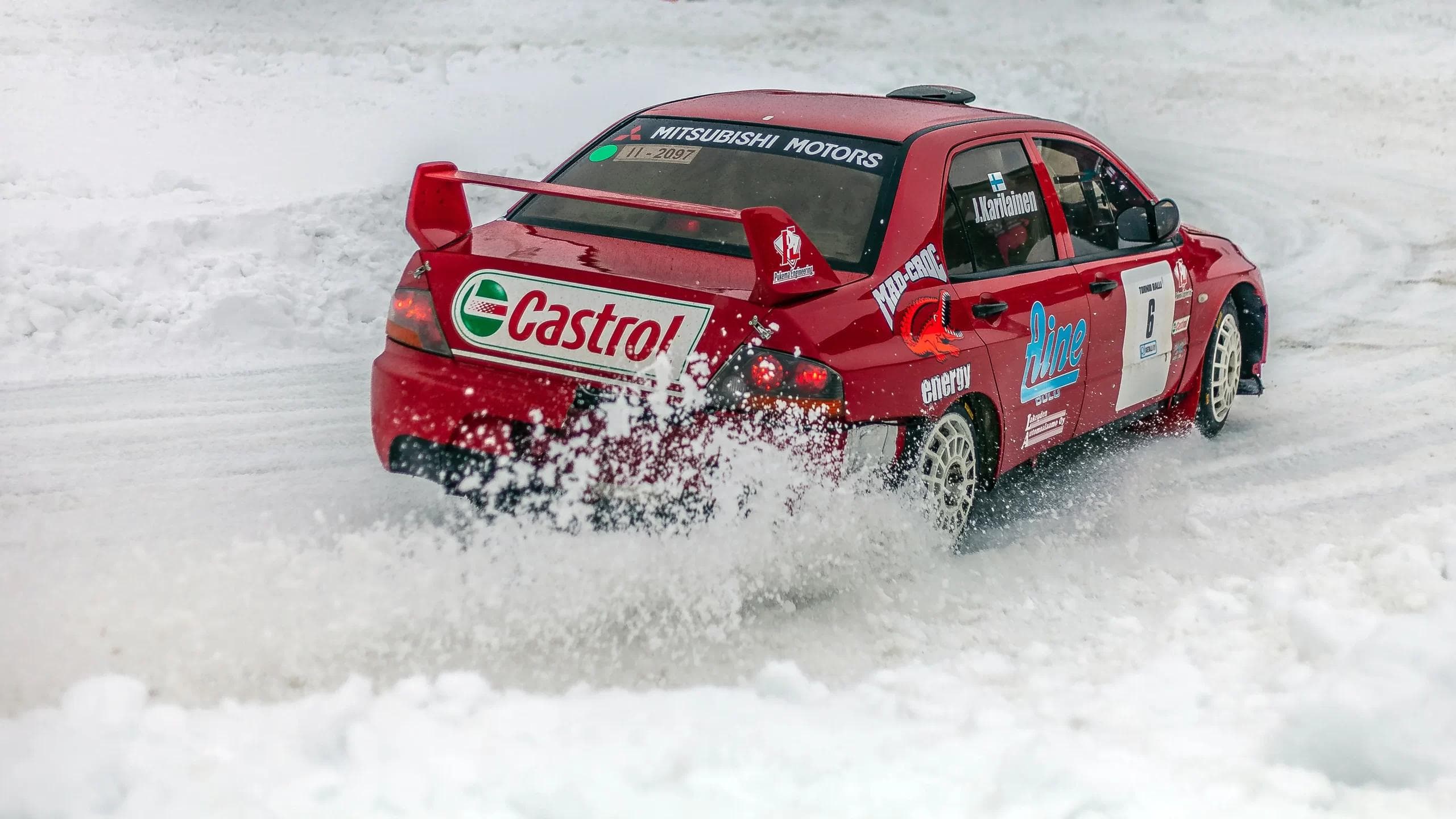 A red Mitsubishi Lancer Evolution rally car with Castrol livery drifting through snow during a winter rally stage, kicking up powder behind the rear wheels.