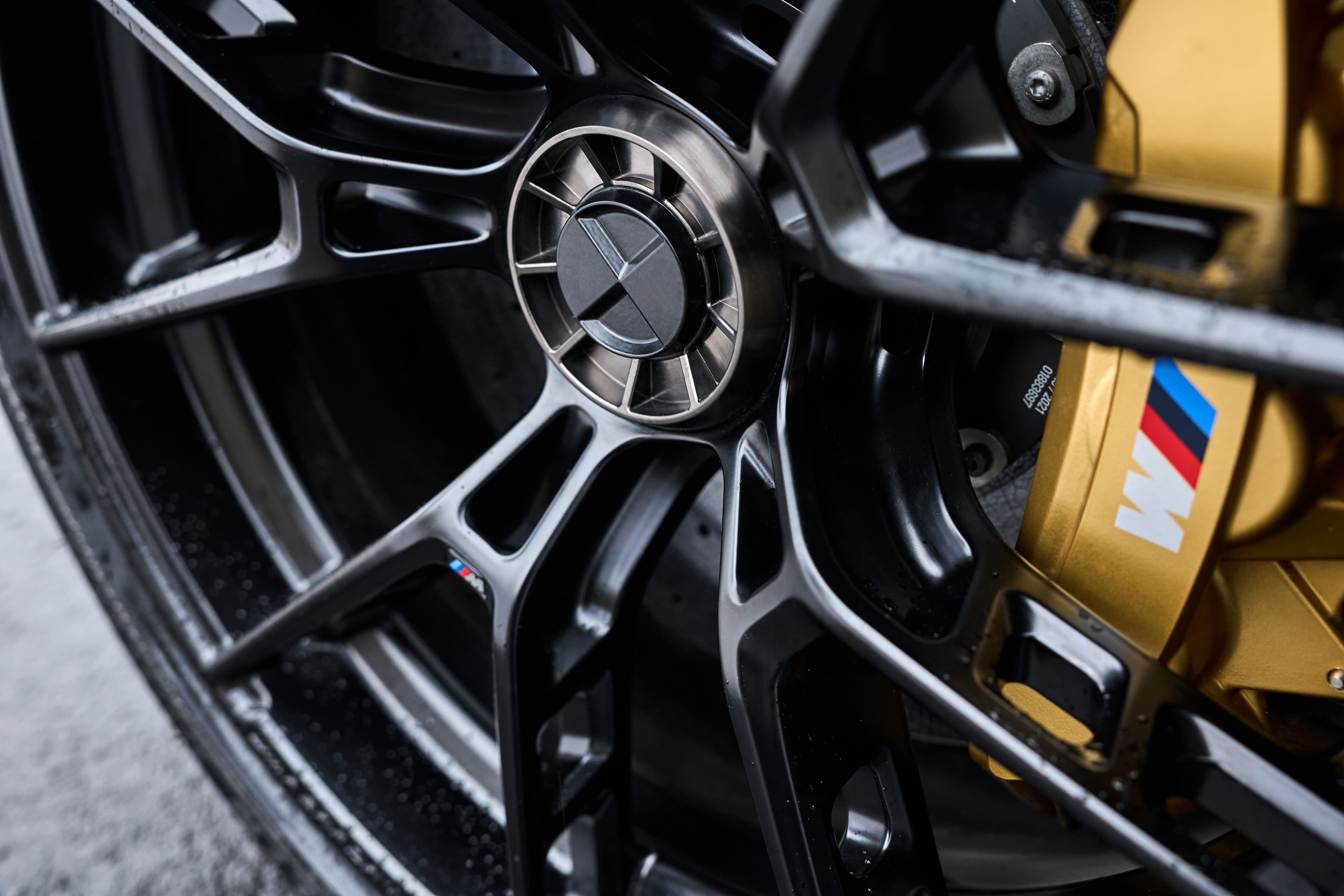 A detailed shot of BMW’s new center-lock wheel, featuring a black multi-spoke design and a gold M Performance brake caliper with the M logo.