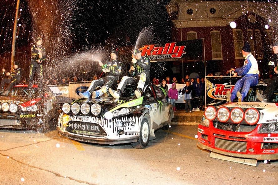 Ken Block and teammates celebrating on rally cars at a Rally America event, spraying champagne in front of a crowd. The Monster Energy and Dirt 2-branded Ford Fiesta is central, flanked by other rally cars with iconic lighting setups.