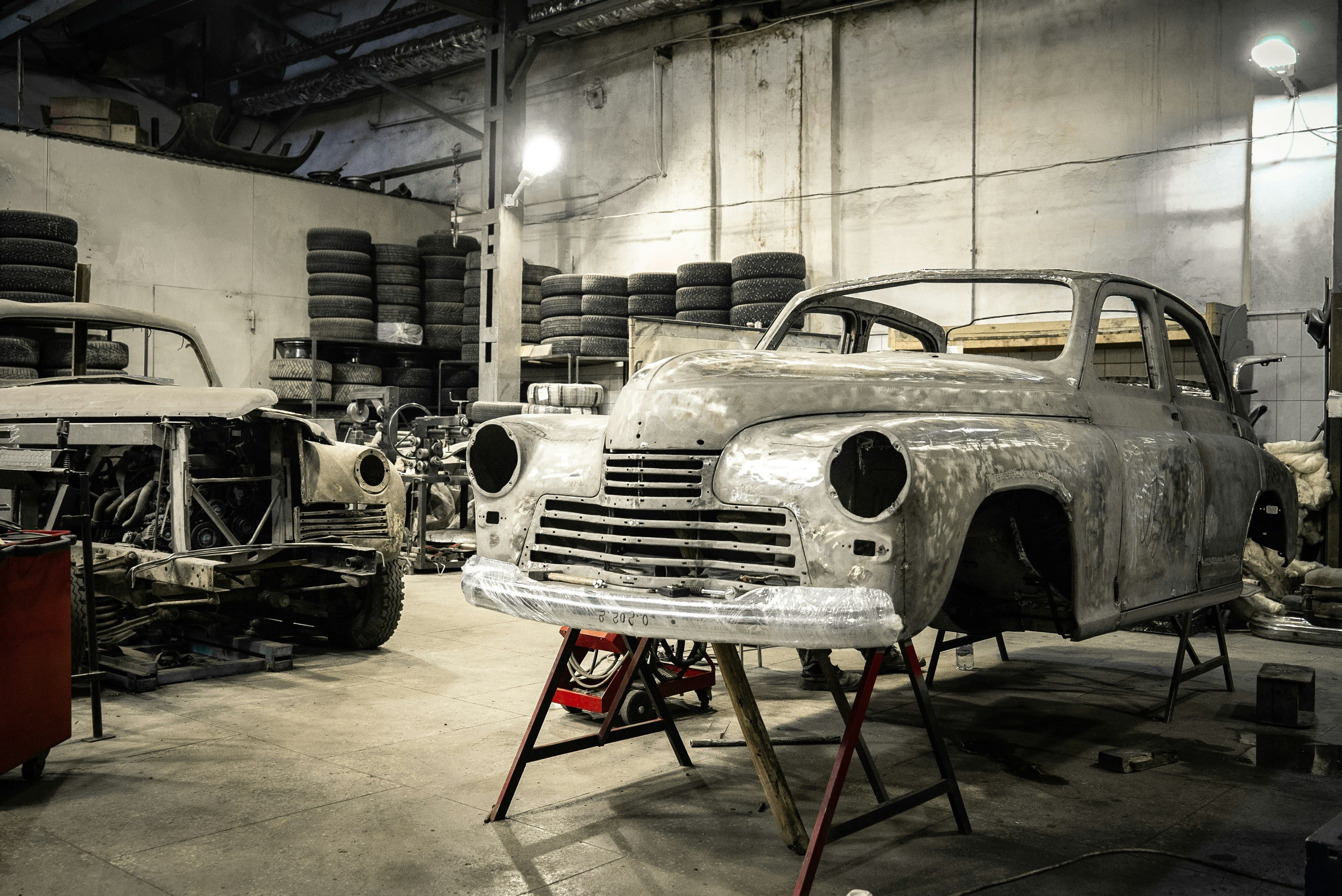 Bare metal classic car shells undergoing restoration in a workshop, with spare tires stacked in the background.