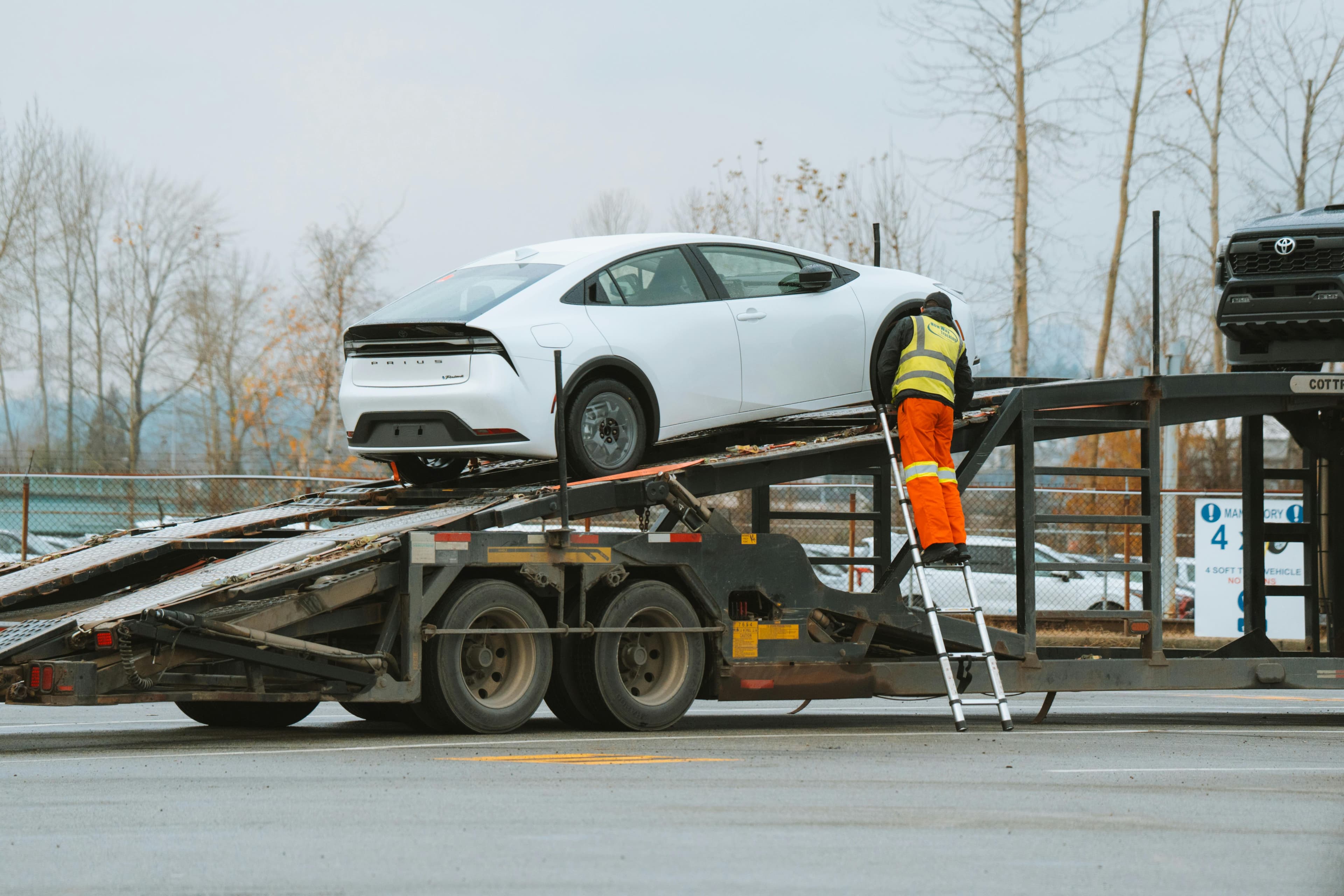 A white Toyota Prius is being loaded onto a car carrier transport truck. A worker in high-visibility clothing and orange safety pants climbs a metal ladder to secure the vehicle. The setting is an outdoor lot with leafless trees in the background, indicating a colder season.
