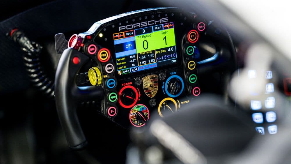Close-up of a Porsche motorsport steering wheel, showing a colourful array of buttons, knobs, and a digital display screen with telemetry and car status information.
