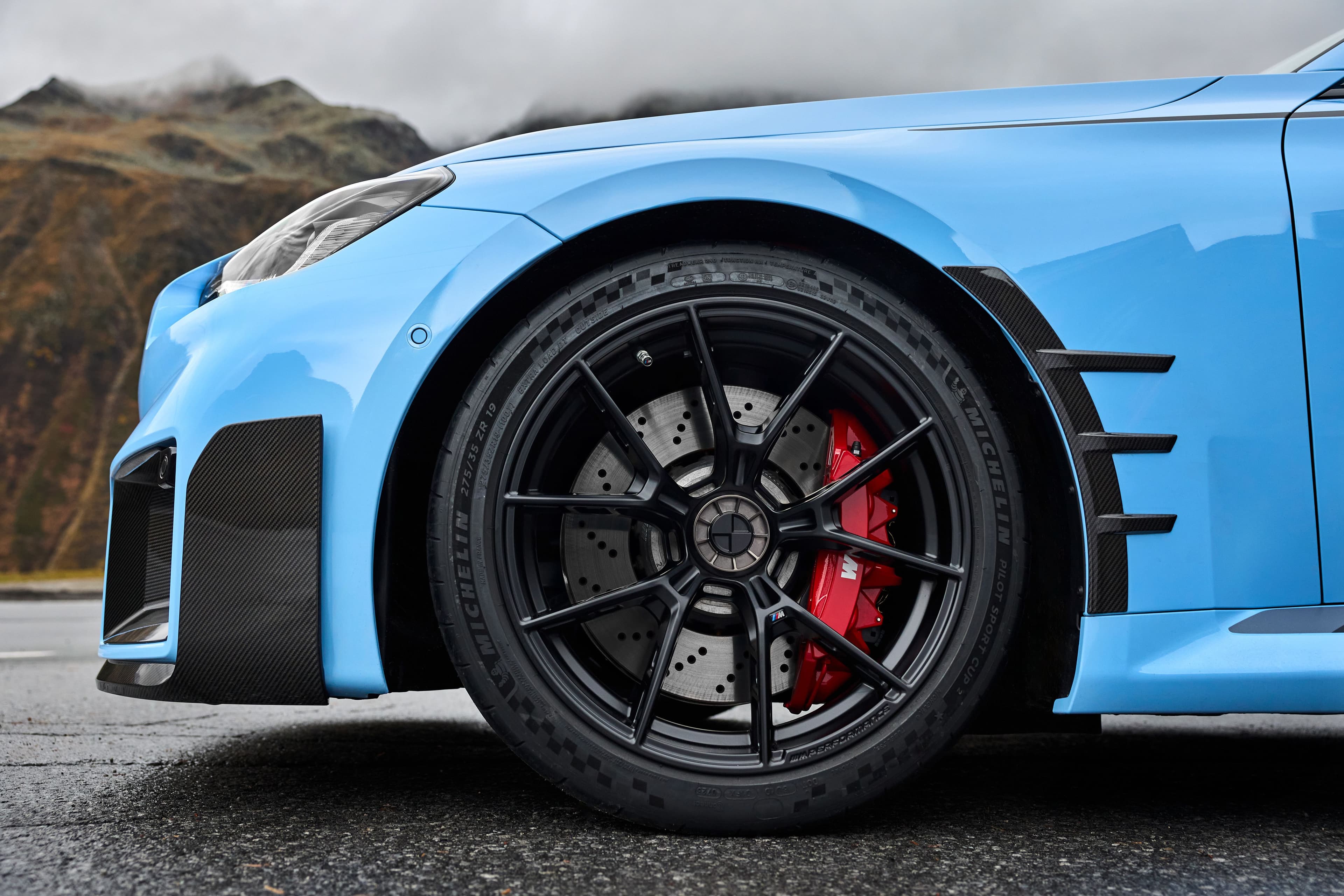 The front wheel of a blue BMW M2, showcasing the center-lock system, red M Performance brake calipers, and Michelin Pilot Sport Cup 2 tires, set against a mountain backdrop.