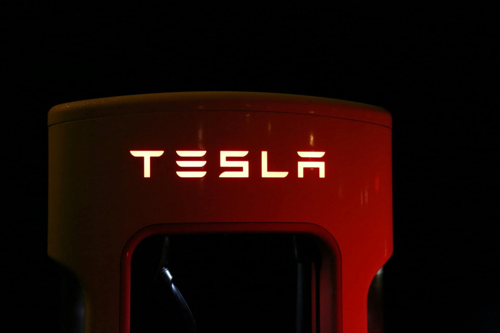 A glowing Tesla logo on a red Supercharger station against a dark nighttime sky.