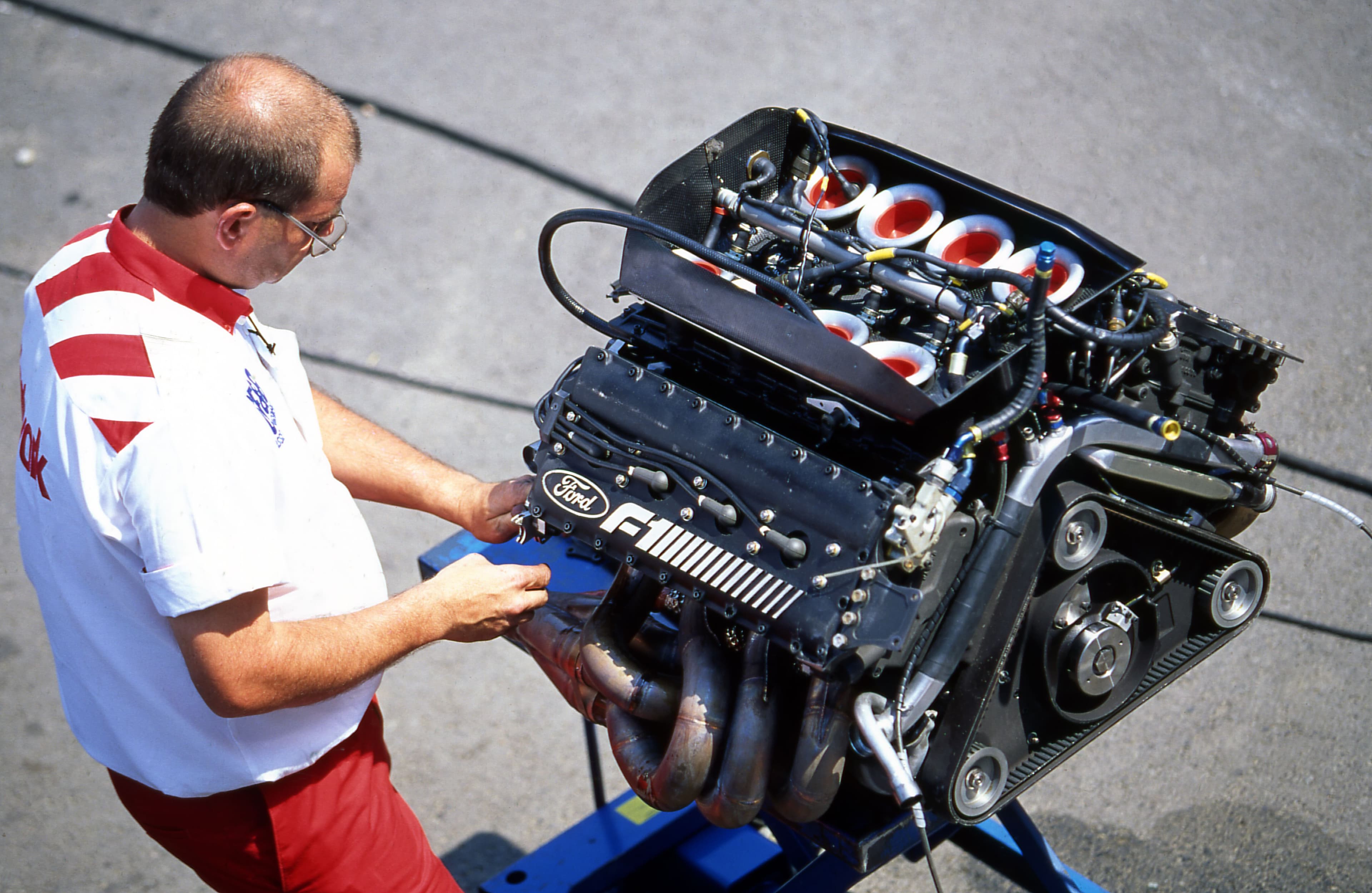 Engineer inspecting a Ford-Cosworth Formula 1 V8 engine at the 1990 Hungarian Grand Prix, featuring individual throttle bodies with red velocity stacks and exposed exhaust headers mounted on a stand in the paddock area.