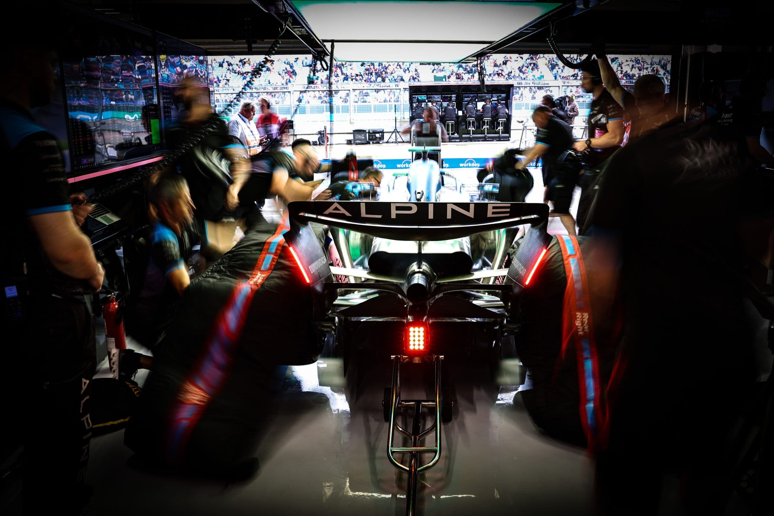 An Alpine Formula 1 car from behind in a pit garage during a race weekend, surrounded by a flurry of mechanics mid-pit stop, with motion blur capturing the intensity.