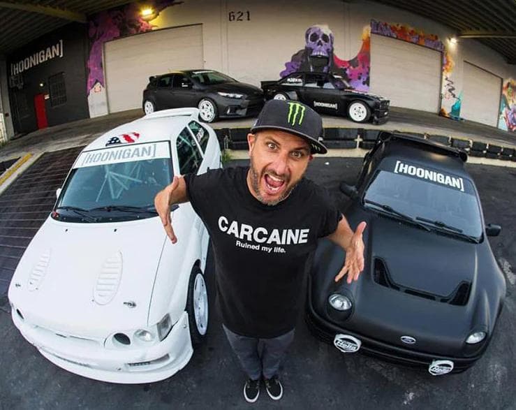 Ken Block making an energetic pose between two Hoonigan-branded cars, a white mk6 Ford Escort and a matte black Ford RS2000, with an MK2 Ford Escort and Ford Focus in the background, outside the Hoonigan headquarters. Wearing a 'Carcaine Ruined My Life' shirt and Monster Energy cap.