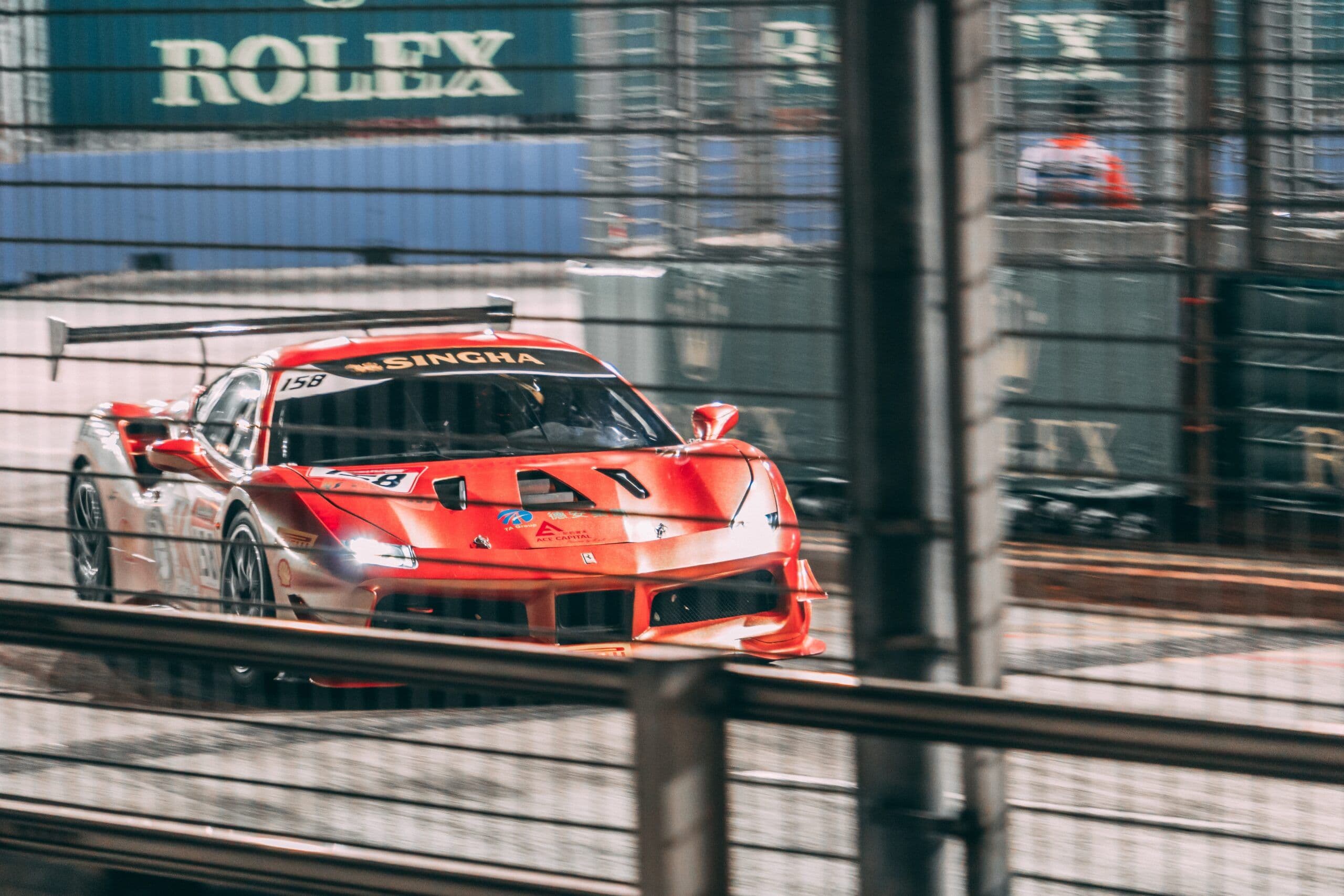 A red Ferrari 488 GT3 race car with Singha sponsorship drives past fencing on a night-time city street circuit, headlights on and bodywork reflecting artificial lights.