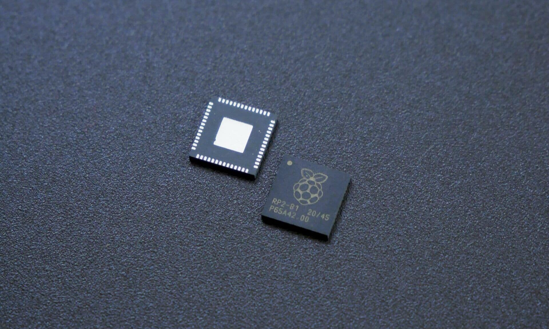 Two Raspberry Pi RP2040 microcontroller chips resting on a dark textured surface, one showing the etched logo and part number.