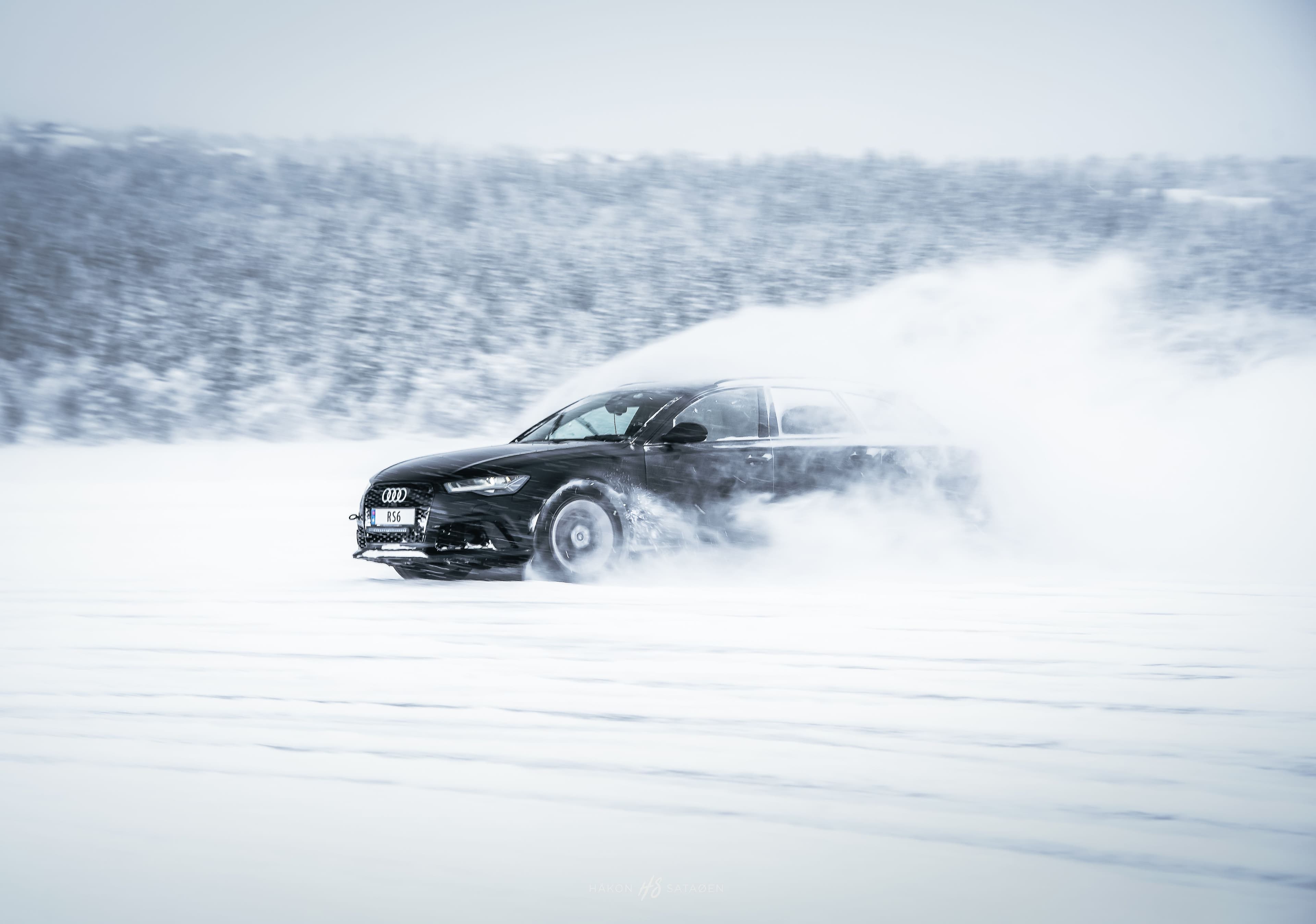 A black Audi RS6 Avant sliding sideways across a snowy landscape, kicking up a large plume of powder as it drifts across a frozen surface in a winter test environment.
