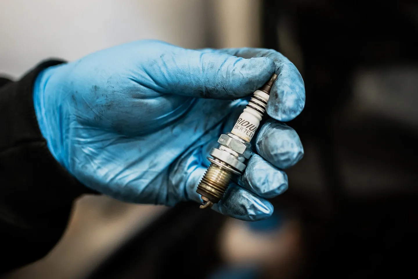 Technician wearing blue gloves holding a used iridium spark plug removed during an engine inspection.
