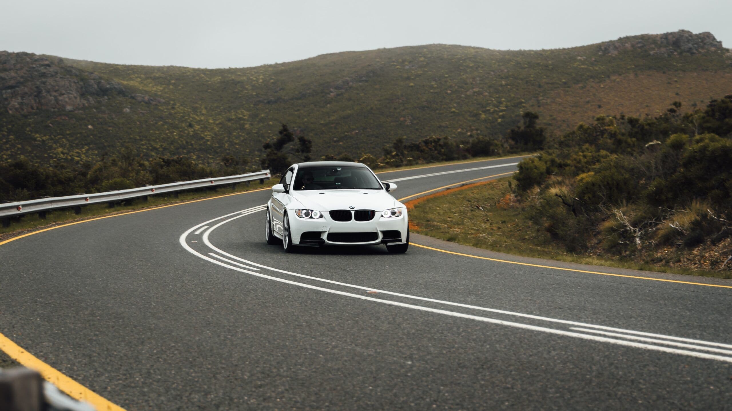 A white BMW E92 M3 navigating a sweeping bend on a scenic mountain road, with overcast skies and green hills in the background.