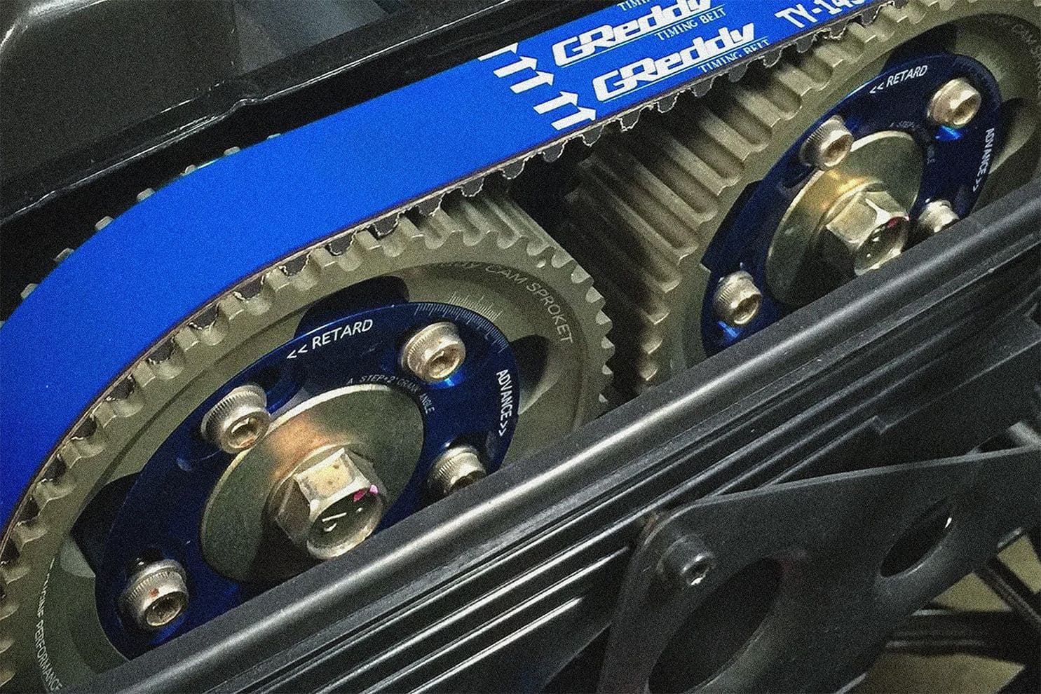 Close-up of GReddy adjustable cam gears and blue timing belt installed on a Toyota 2JZ or 1JZ engine, showing advanced and retard timing markings.