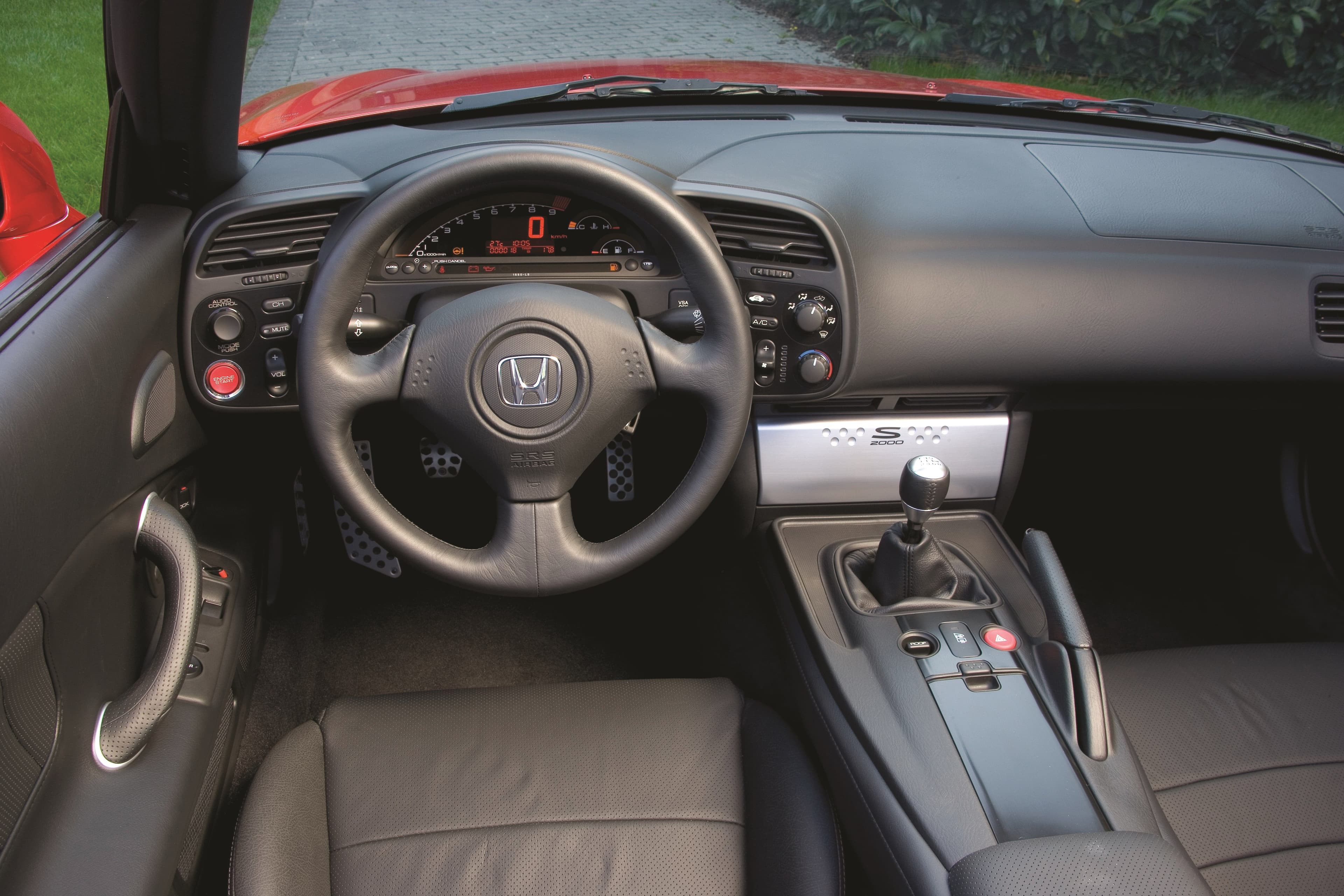 Interior of the Honda S2000, highlighting the leather steering wheel, digital instrument cluster, and 6-speed manual gear shifter in a driver-focused cockpit design.