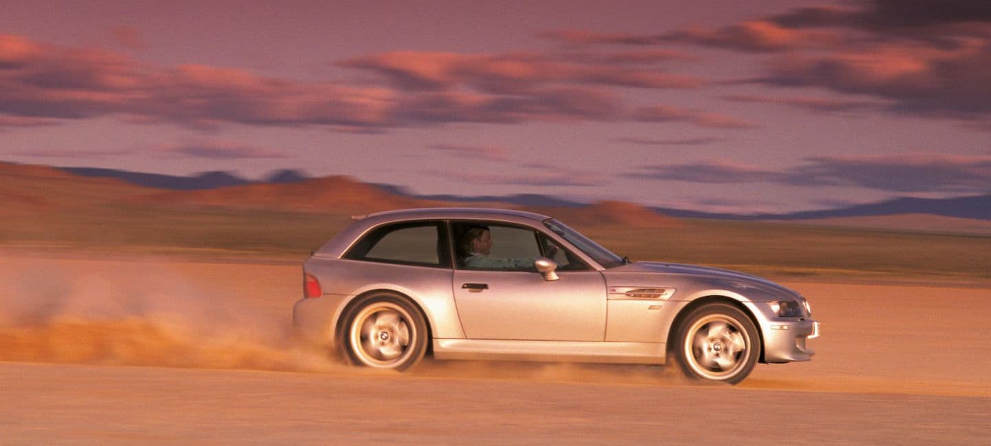 Silver BMW Z3 M Coupe drifting through a dry lake bed at speed, kicking up a trail of dust under a colourful sunset sky.