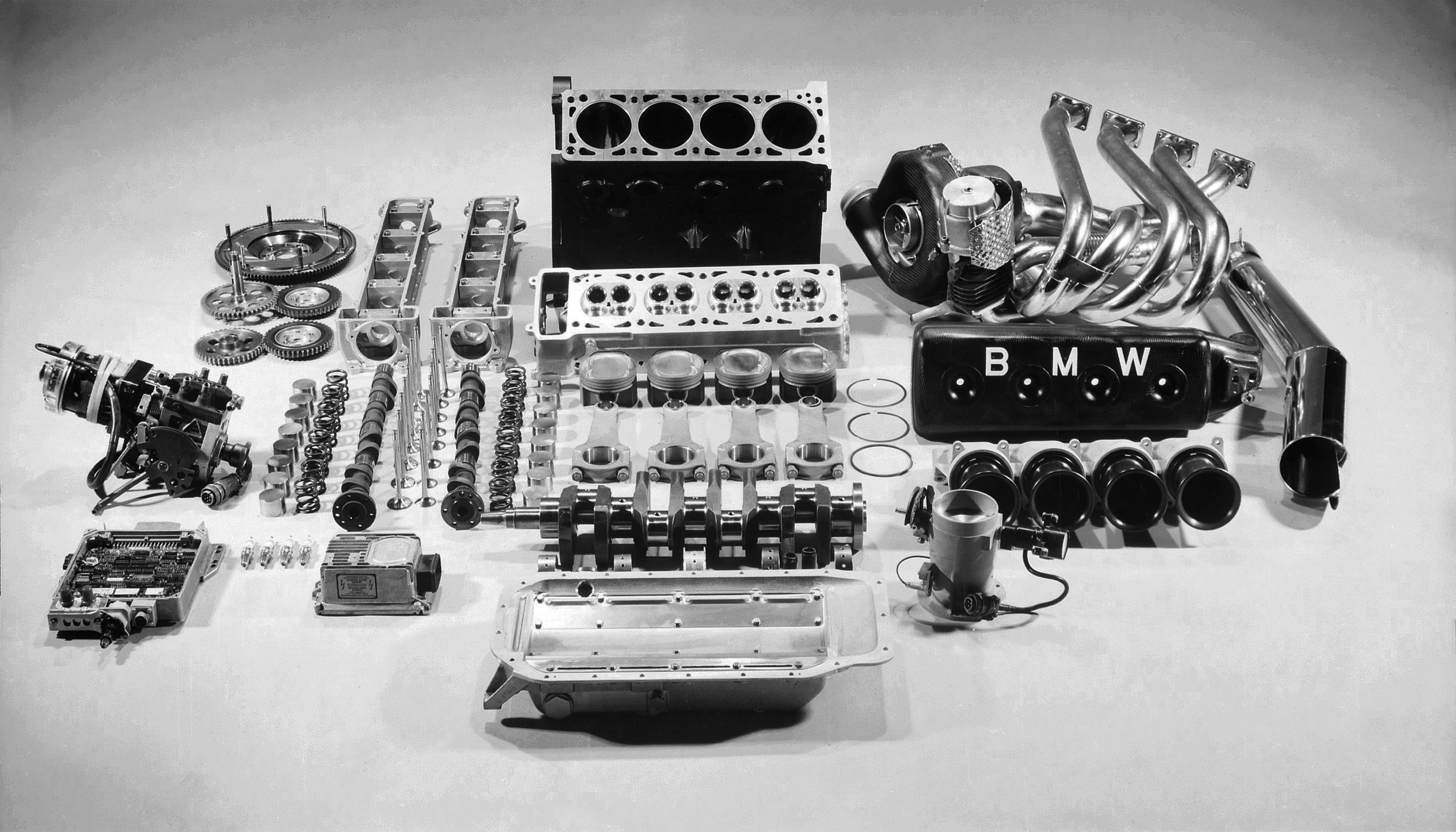 BMW Formula 1 engine from 1983 fully disassembled and neatly laid out, showcasing all internal components including pistons, crankshaft, camshafts, turbocharger, cylinder head, and exhaust manifold, with the BMW-branded valve cover prominently displayed.