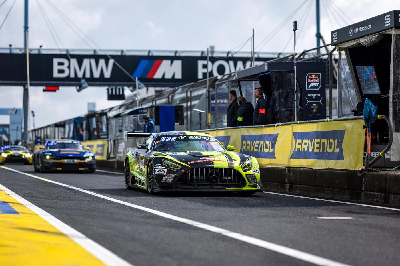 A black and neon yellow Mercedes-AMG GT3 leaving the pit lane during the Nürburgring 24 Hours, with Ravenol pit wall branding and BMW M Power gantry overhead, GT cars queued in the background